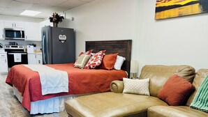 A hotel room with a bed, a sofa, a kitchenette, and a painting on the wall.