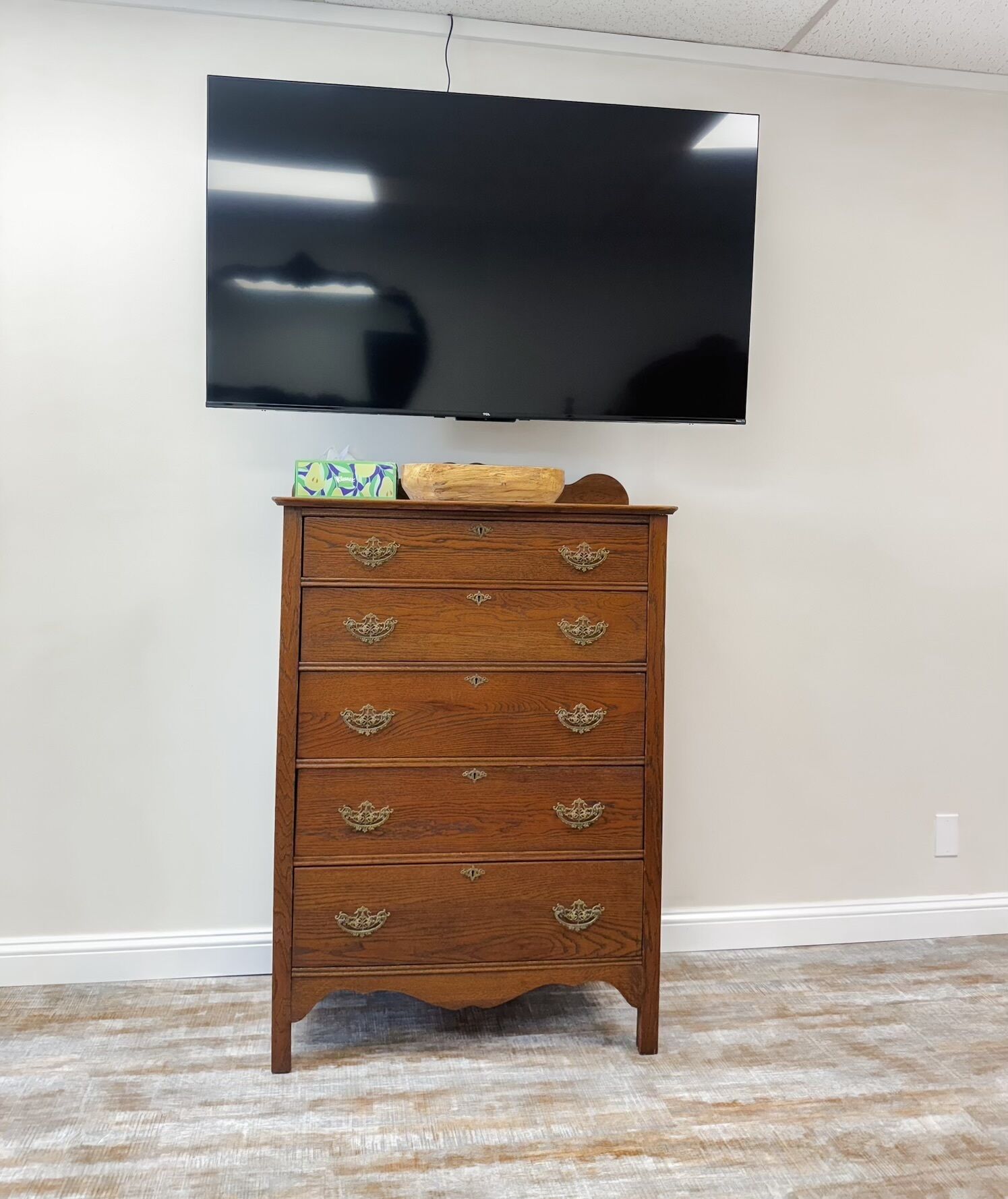 A wooden dresser with ornate handles against a plain wall, a flat-screen TV mounted above it, and a carpeted floor.