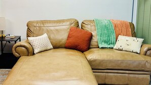 A tan leather sofa with a matching ottoman, a green and orange throw blanket, and a wall-mounted painting of a sunset.