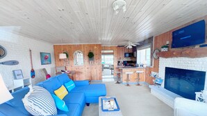 A modern living room with a blue sofa, a coffee table, and a fireplace. There is a kitchen area with a bar and a television mounted on the wall.