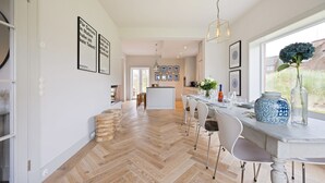 A spacious kitchen with a herringbone floor pattern, a dining area with a white table and chairs, and a large window with a view of greenery. - Dünen.Way, Germany (List auf Sylt)