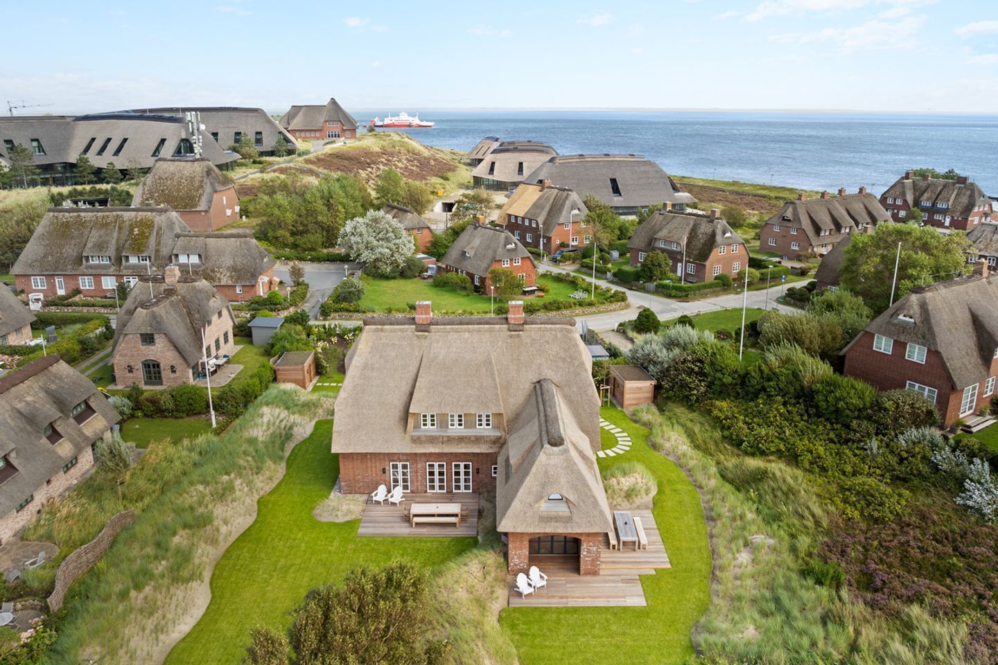 An aerial view of a coastal village with traditional thatched-roof houses, a central building with a large deck, and a ship in the background.