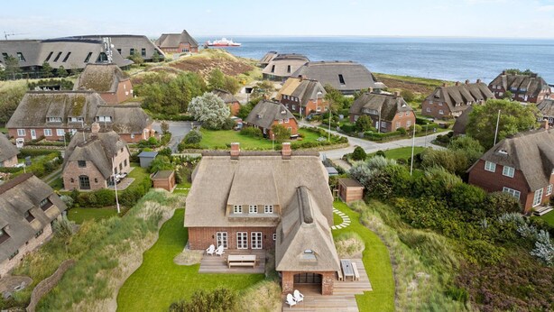 An aerial view of a coastal village with traditional thatched-roof houses, a central building with a large deck, and a ship in the background.