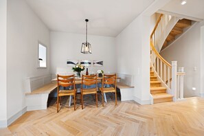 A dining area with a wooden table and chairs, a hanging light fixture, and a staircase with wooden steps and white railings. - Heide.Way, Germany (List auf Sylt)