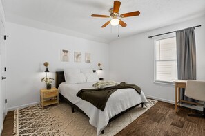 Ein Schlafzimmer mit einem Bett, Nachttischen, einem Deckenventilator, einem Fenster mit Vorhängen und einem Sessel.
