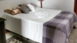Een net opgemaakt bed met een wit-paars geruit deken, een houten hoofdbord en een klein houten plankje erboven met een tas met luipaardprint en een paar schoenen.