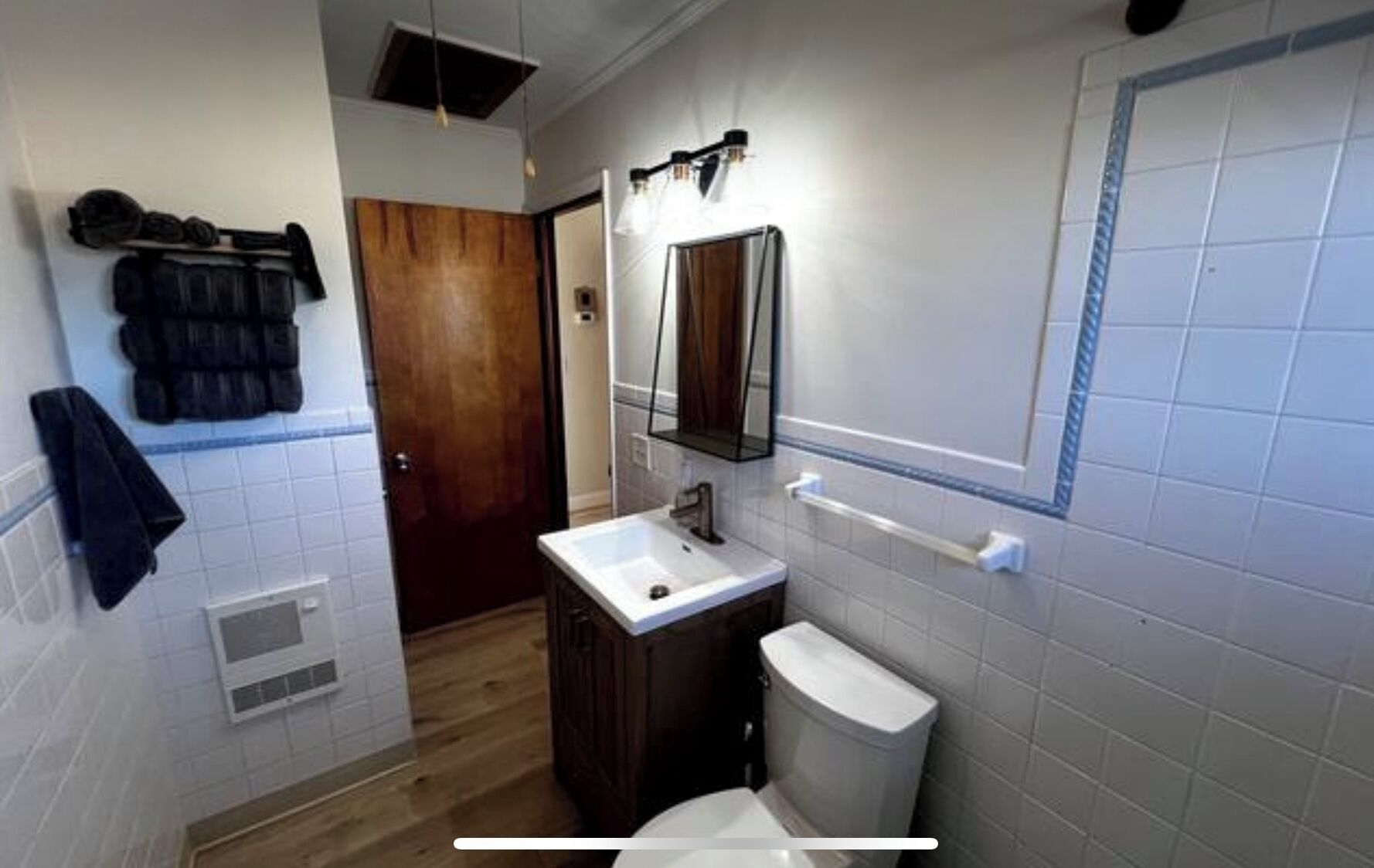 Un bany amb un lavabo blanc, un armari de fusta fosca i un vàter blanc. Hi ha un suport de tovalloles a la paret i un mirall sobre el lavabo.
