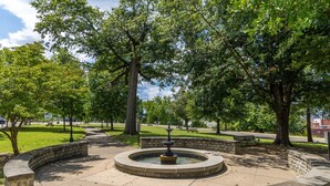 Een ronde stenen fontein omringd door een gebogen stenen bank in een park met bomen en een picknickplaats.