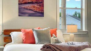 Een net opgemaakt bed met een witte dekbedovertrek en meerdere kussens in verschillende kleuren. Een raam met uitzicht op een huis en bomen is zichtbaar, en een landschap schilderij hangt aan de muur.