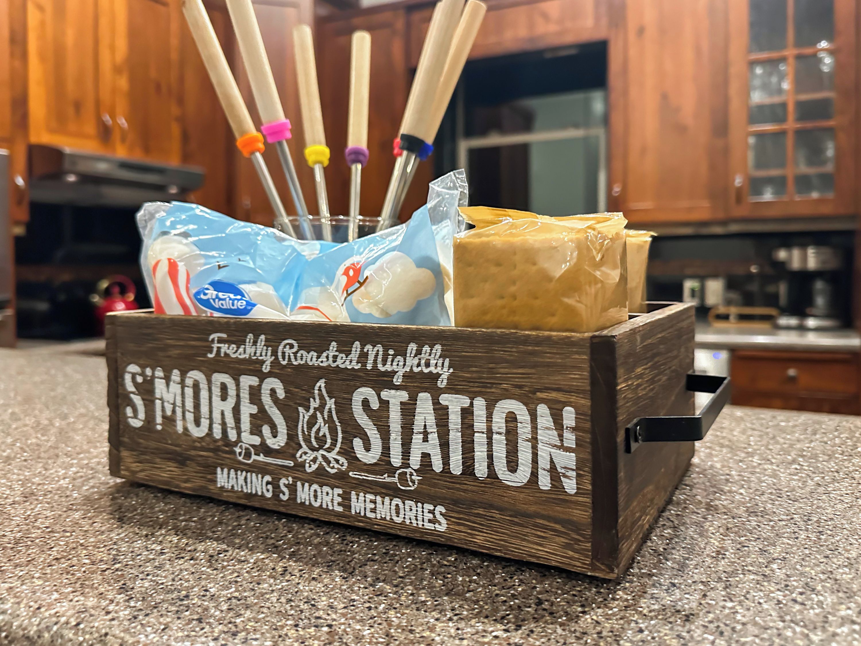 Une boîte en bois portant l’inscription « Station de préparation de s’mores fraîchement grillés tous les soirs », contenant un sachet de guimauves et une tablette de chocolat, ainsi que plusieurs brochettes colorées pour les guimauves.