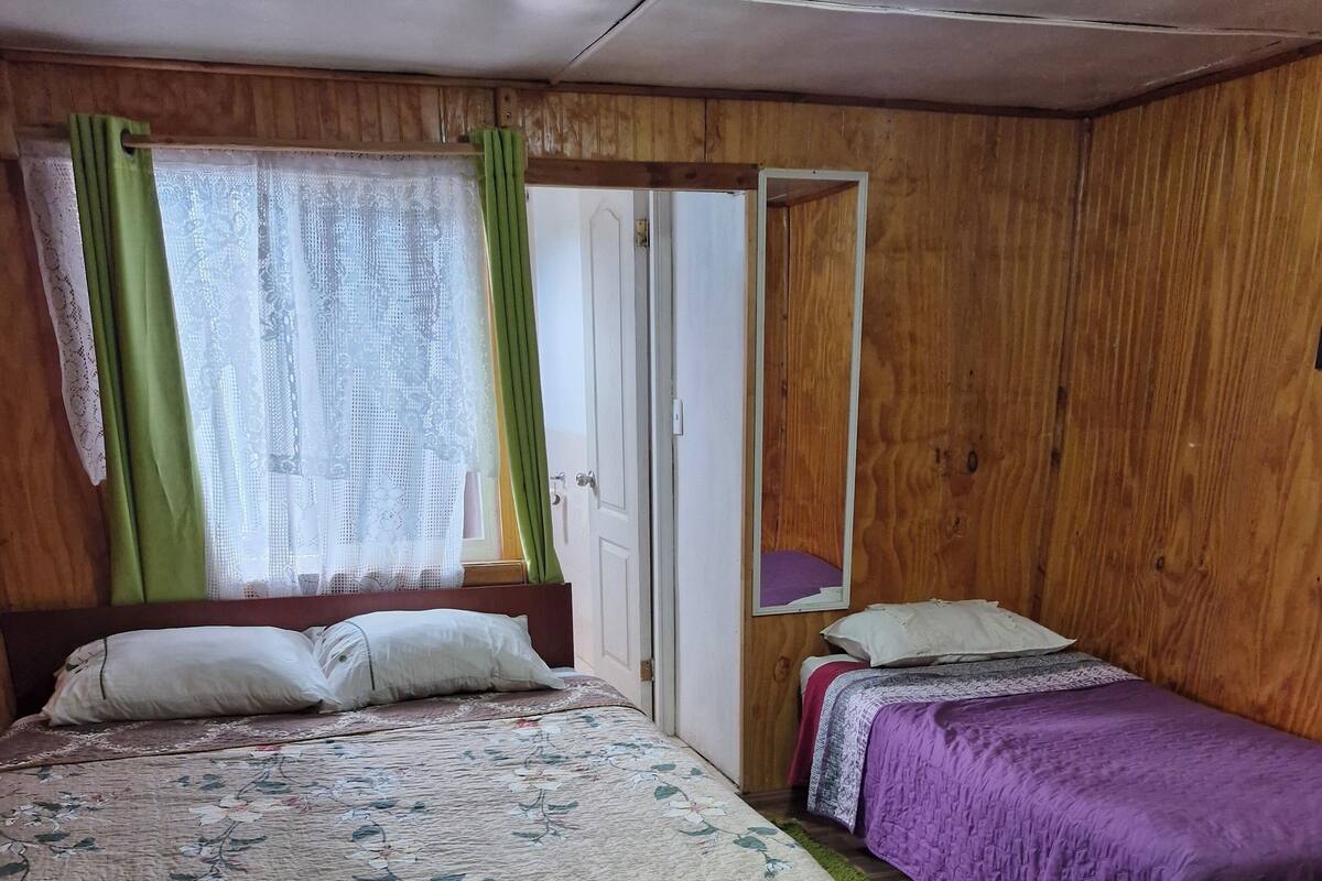 Una habitación con una cama grande, una cama más pequeña, paredes de madera, una puerta y una ventana con cortinas.