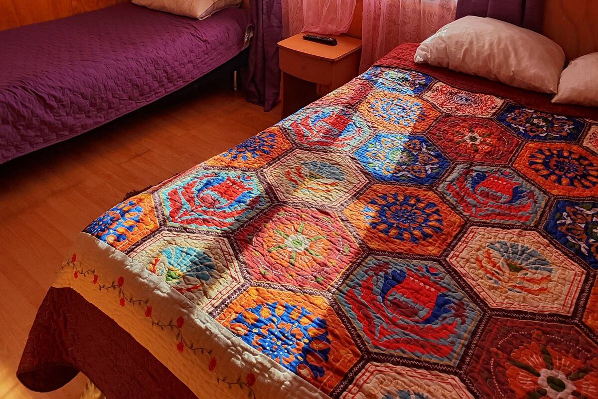 Habitación con dos camas, una con cubrecama morado y la otra con colcha multicolor. Se observa un cabecero de madera, una mesita con control remoto y una ventana con cortinas moradas.