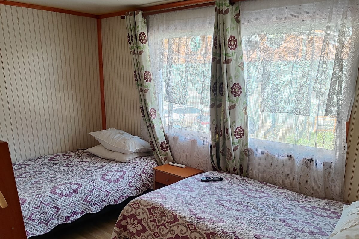 Una habitación con dos camas, suelo de madera, una ventana con cortinas y una lámpara en el techo.