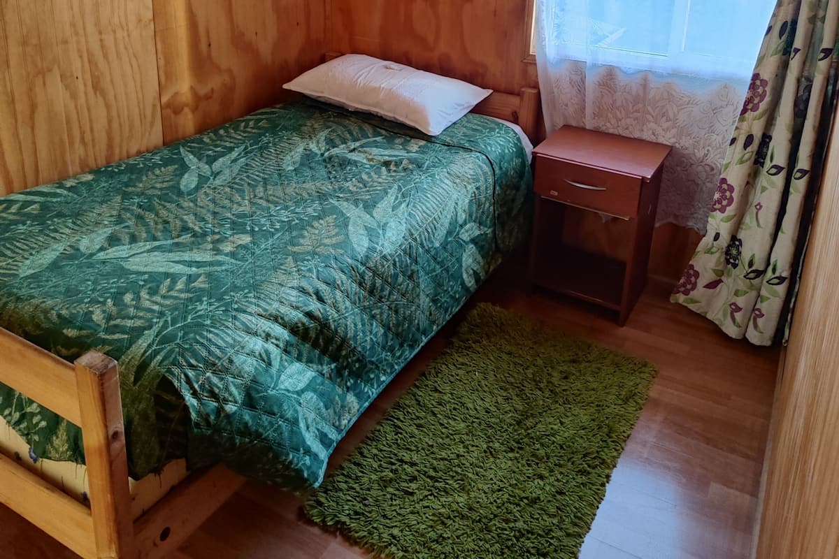 Una habitación pequeña y acogedora con paredes de madera, una cama individual con colcha verde estampada, una mesita de noche de madera, un tapete verde y una ventana con cortina floral.