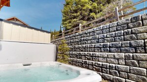 A hot tub next to a stone wall with a wooden fence and a building in the background. - Beautiful Echo Landing Retreat, Sleeps 8, 3 Bdrm, Private Hot Tub, Ski In/Out  (Sun Peaks)