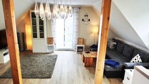A spacious loft apartment with a high ceiling, exposed wooden beams, a large window, a cozy seating area with a sofa and armchairs, and a dining table. - Vacation home directly on the dune, approx. 50 m to the Baltic Sea - Markgrafenheide (Rostock)