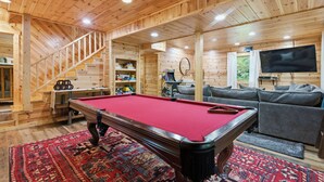 A wooden interior with a pool table, sofa, and staircase.