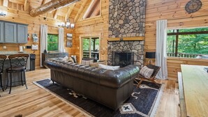 A log cabin interior with a stone fireplace, wooden ceiling beams, a leather sofa, and a dining area.