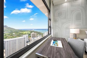Quarto de hotel com uma grande janela com vista para a cidade, uma escrivaninha de madeira, uma poltrona cinza e uma parede com padrão.