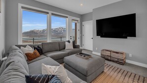 Un salon moderne avec un canapé d’angle gris, un téléviseur à écran plat fixé au mur et une grande fenêtre donnant sur une vue imprenable sur les montagnes.