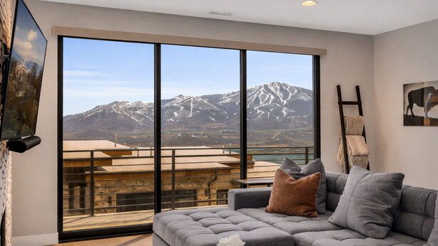 Un salon moderne avec un canapé d’angle gris, un téléviseur à écran plat fixé au mur et une grande fenêtre donnant sur une vue imprenable sur les montagnes.