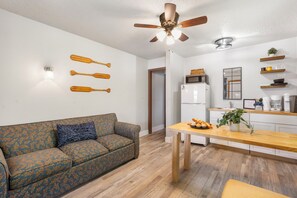 A compact living space with a sofa, a dining table, a ceiling fan, and a kitchen area with a refrigerator and microwave.