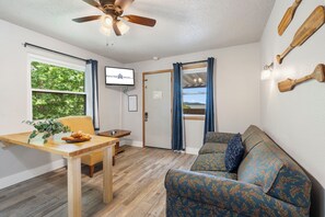 Quarto mobiliado com mesa de madeira, sofá, ventilador de teto e televisão fixada na parede.