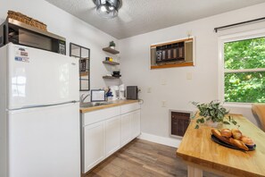 Une cuisine avec des meubles blancs, un four à micro-ondes, un réfrigérateur, une machine à café et une fenêtre donnant sur des arbres.