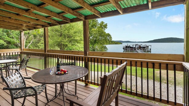A covered outdoor deck with a table and chairs, overlooking a lake and dock.