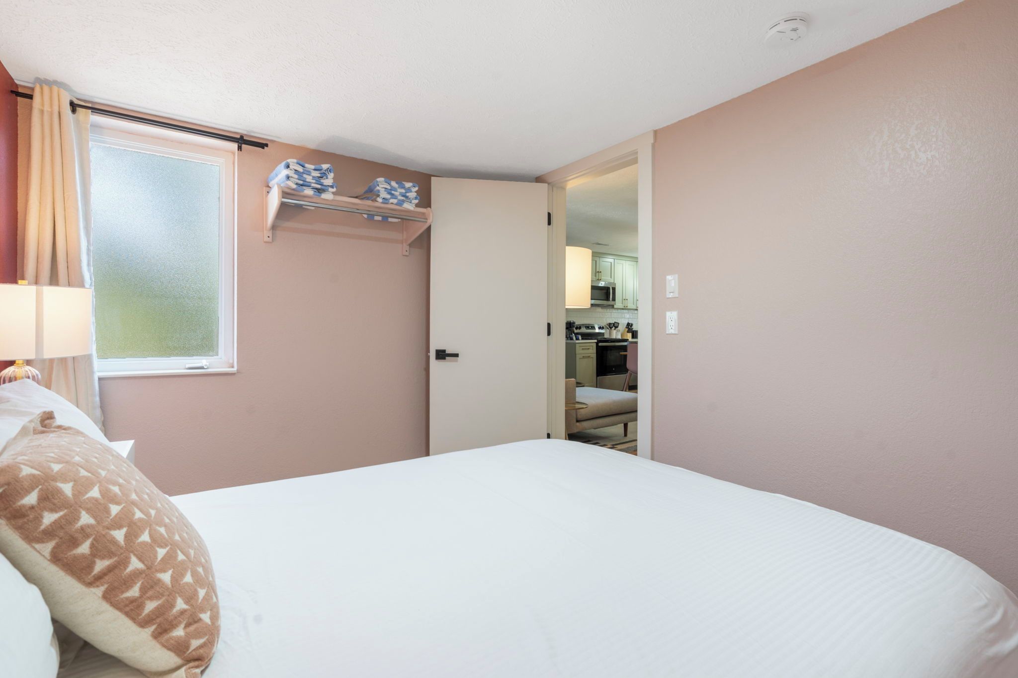 A bedroom with a bed, a window with curtains, a bedside lamp, and a view into a kitchen area.