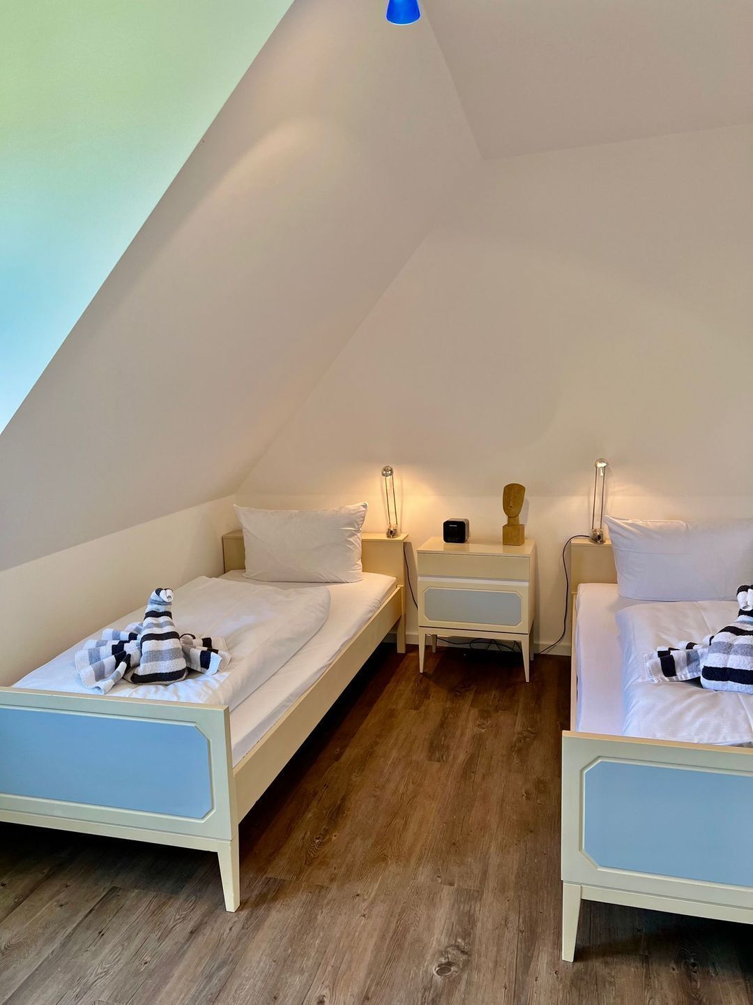 Une petite chambre mansardée, chaleureuse et accueillante, avec deux lits simples, une table de chevet, une lampe et une petite table.