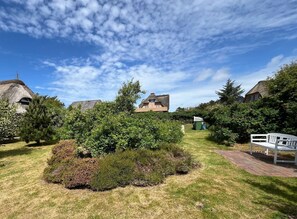 A rural setting with traditional thatched-roof cottages, a well-maintained garden with various shrubs, and a clear blue sky.