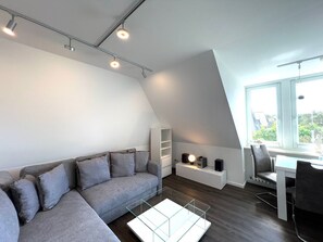 A modern loft apartment with a grey sofa, a glass coffee table, a dining area with a chair, and a white shelving unit.