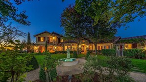 A large, well-lit house with a fountain in the garden, surrounded by trees and a clear sky. - Spacious vineyard estate in Napa Valley with home, guest home, pool, pano views (Calistoga)
