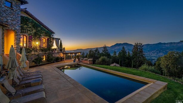 A poolside area with lounge chairs and umbrellas, a stone building with warm lighting, and a view of mountains in the background. - Spacious vineyard estate in Napa Valley with home, guest home, pool, pano views (Calistoga)