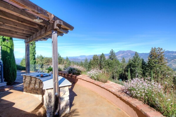 Un patio cubierto con un banco de piedra, una pérgola y vistas a montañas y árboles.