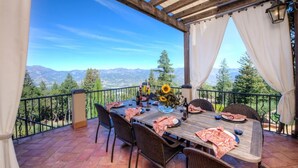 An outdoor patio with a wooden table set for dining, surrounded by wicker chairs, and overlooking a mountainous landscape. - Spacious vineyard estate in Napa Valley with home, guest home, pool, pano views (Calistoga)