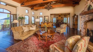A spacious living room with a high ceiling, wooden beams, a fireplace, and a large window overlooking a scenic view. - Spacious vineyard estate in Napa Valley with home, guest home, pool, pano views (Calistoga)