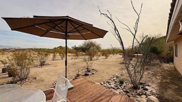 Una terraza de madera con una silla de metal y un paraguas grande, rodeada de vegetación desértica y un edificio.