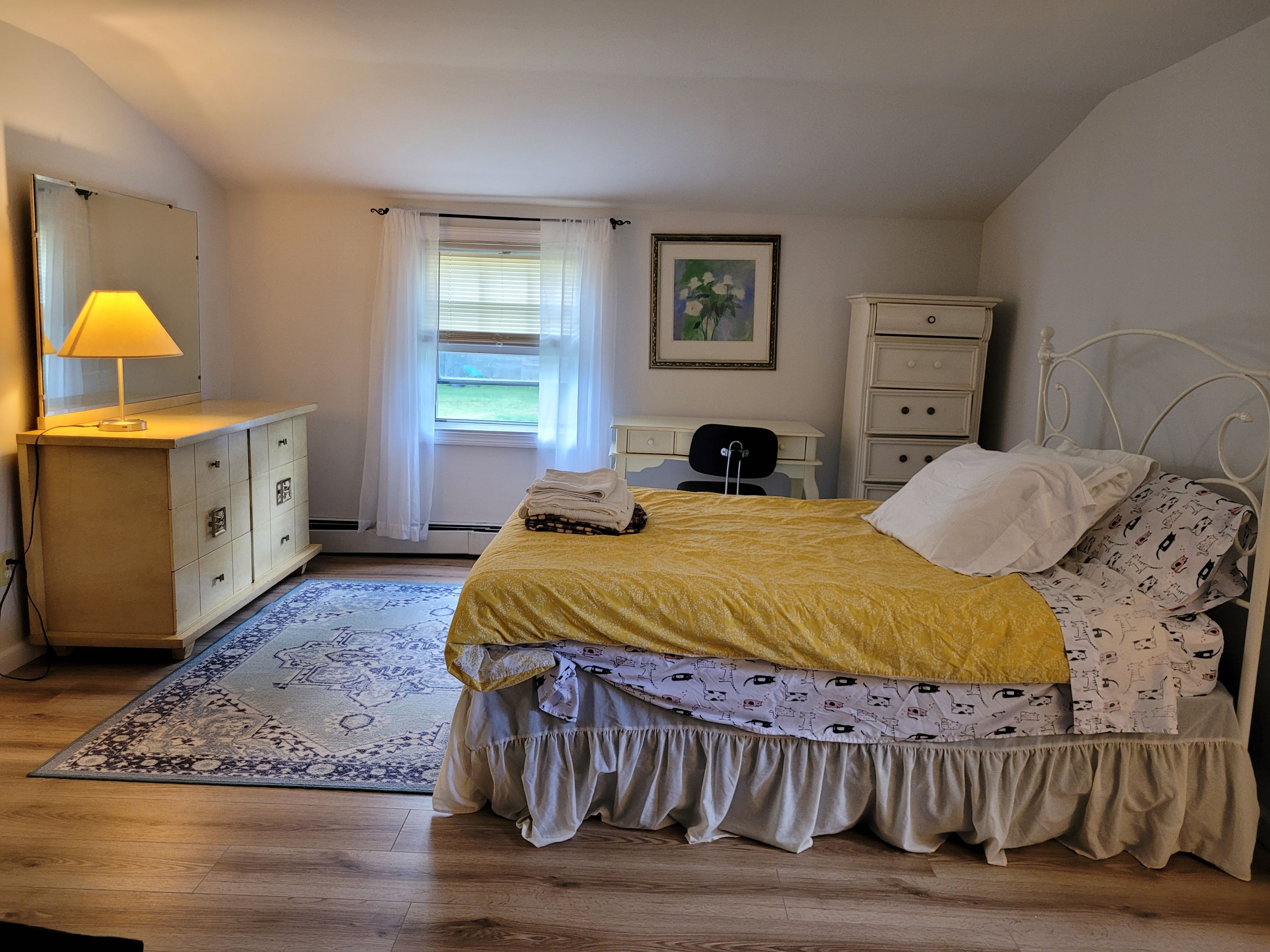 A bedroom with a bed, a dresser, a desk, a chair, a lamp, and a window with curtains.