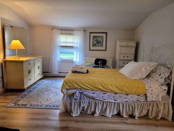 Une chambre à coucher comprenant un lit, une commode, un bureau, une chaise, une lampe et une fenêtre avec des rideaux.