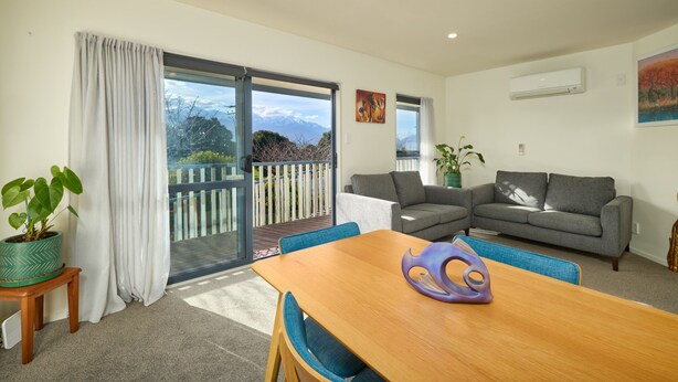 A modern living room with a wooden dining table, two grey sofas, and a balcony with a view of mountains. - Handy Location | Ocean & Mountain Views | Spacious Yard - KK46451 (Kaikoura)