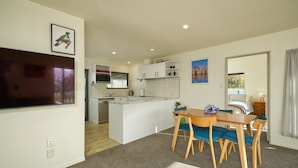 A modern kitchen with white cabinets, a central island, and a dining area with a table and chairs. - Handy Location | Ocean & Mountain Views | Spacious Yard - KK46451 (Kaikoura)