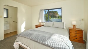 A bedroom with a large bed, two bedside tables with lamps, a window with a view of greenery, and a door leading to a bathroom. - Handy Location | Ocean & Mountain Views | Spacious Yard - KK46451 (Kaikoura)