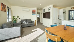 A modern living room with a grey sofa, a wooden dining table with blue chairs, and a kitchen area with white cabinets and a clock. - Handy Location | Ocean & Mountain Views | Spacious Yard - KK46451 (Kaikoura)