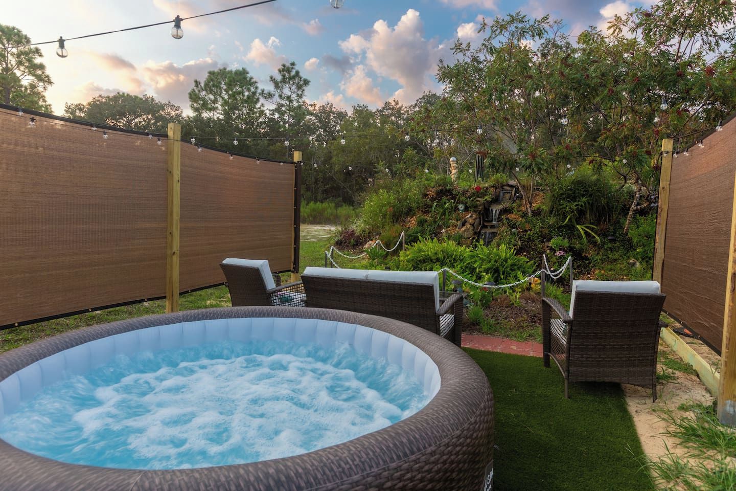 A hot tub with wicker chairs, surrounded by a garden with a fence and string lights.
