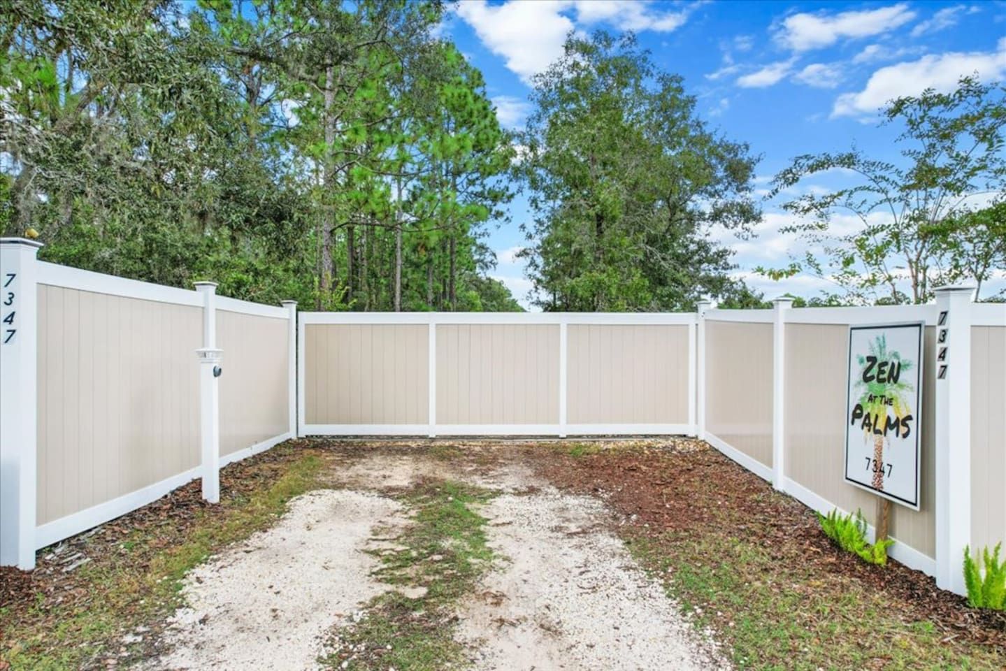 A fenced backyard with a gravel path, trees, and a sign that reads 'ZEN AT THE PALMS 7347'.