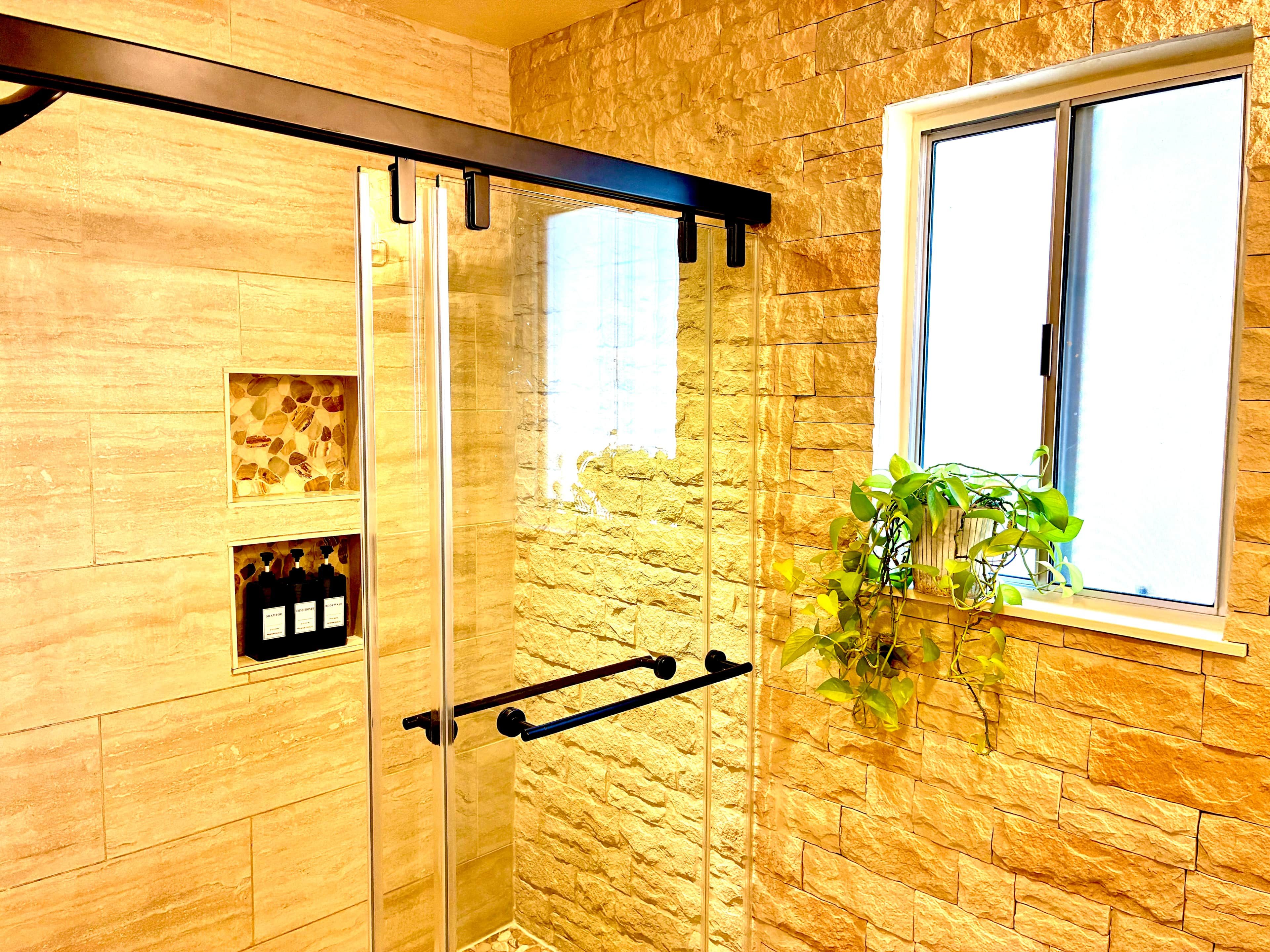 A bathroom with a glass shower door, a window with a plant, and a shelf with bottles.