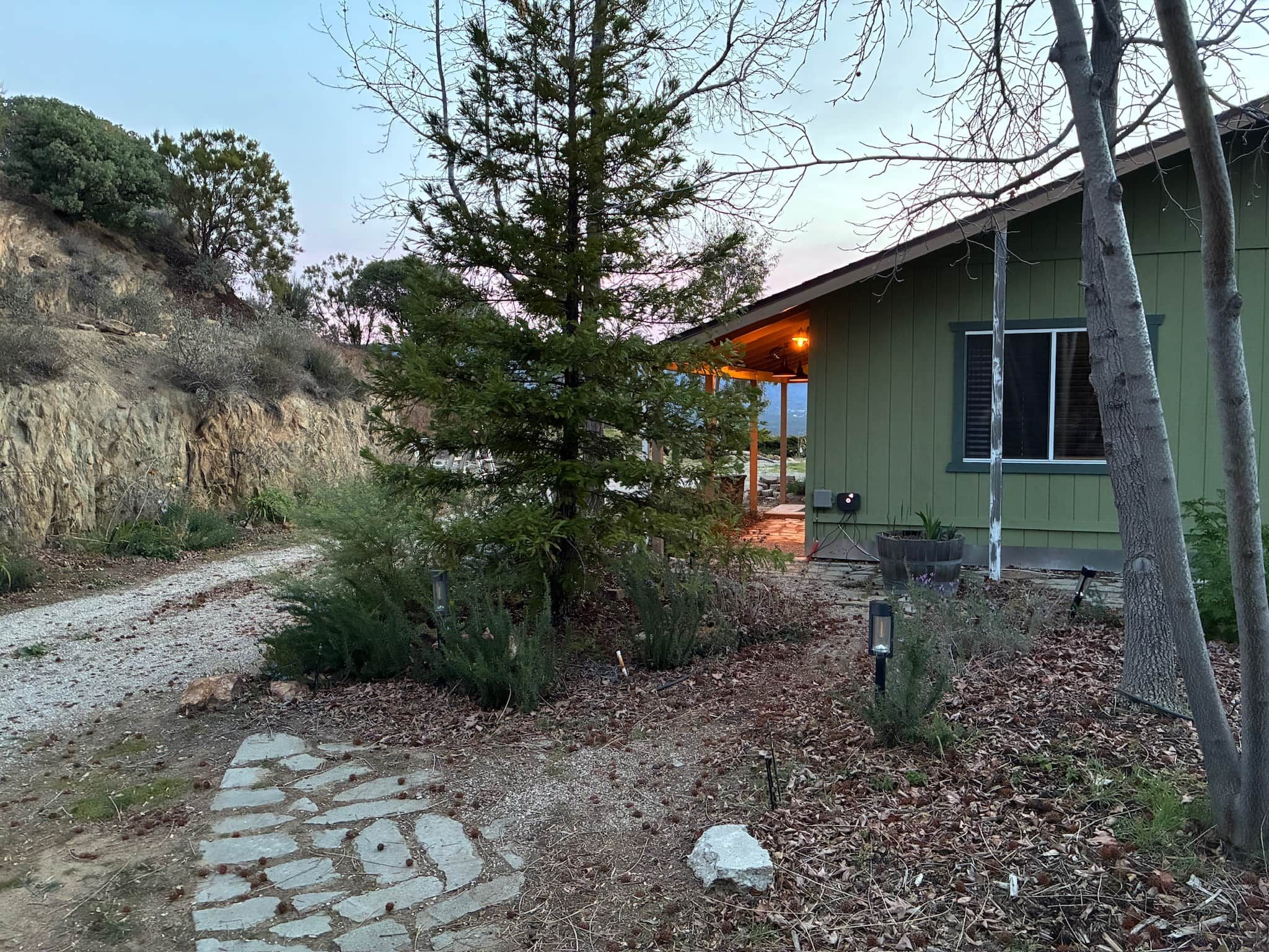 A house with a stone pathway leading to a wooden door, surrounded by trees and a hillside.