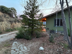 Una casa con un camino de piedra que lleva a una puerta de madera, rodeada de árboles y una ladera.